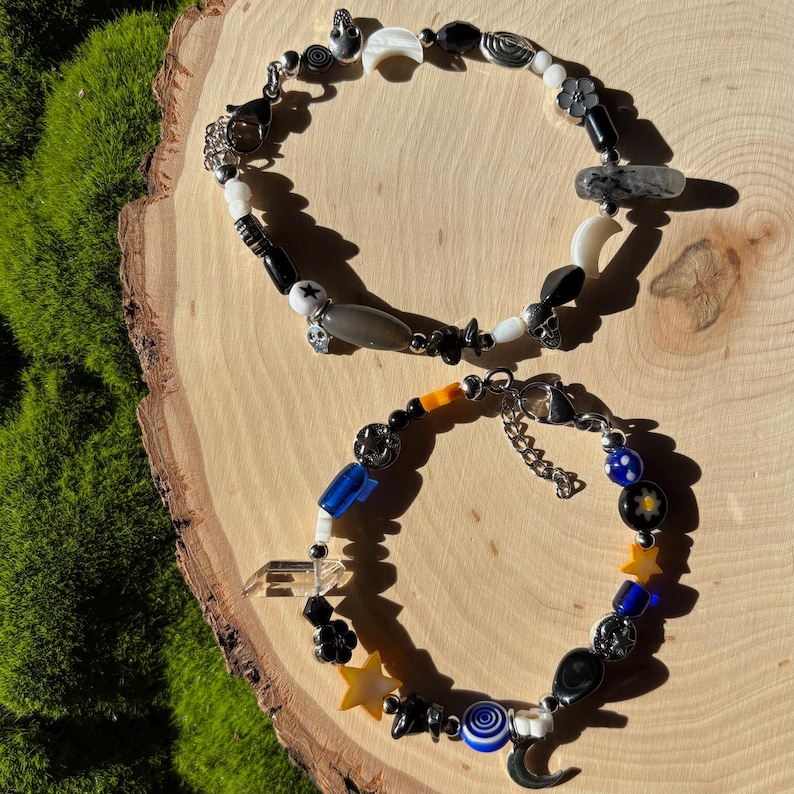 K&ouml;nnte beinhalten: Zwei Armb&auml;nder mit Perlen mit himmlischen und abstrakten Designs. Ein Armband hat schwarze, wei&szlig;e und graue Perlen mit Mond- und Sternanh&auml;ngern. Das andere enth&auml;lt blaue, gelbe und schwarze Perlen mit Stern- und Mondanh&auml;ngern.