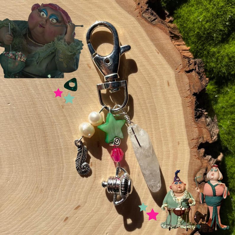 May include: A silver keychain with a lobster clasp, adorned with a feather charm, pearls, a green star, pink beads, a clear crystal, and a silver decorative element. The background features a wooden surface and green moss.
