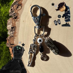 May include: Silver-toned keychain with a lobster clasp, featuring a skeleton charm, a white bead with a black star, and a dark stone. Additional charms include a black bead and a silver spiral. The background includes a wooden surface and a green mossy area.
