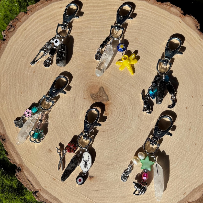 May include: Six silver-toned keychains with various charms, including crystals, a yellow dragonfly, flowers, and a spider. The keychains are displayed on a light brown, textured wooden surface.