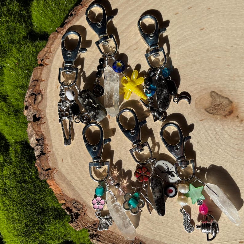May include: A collection of silver keychains with various charms, including a skeleton, crystals, a yellow dragonfly, and a spider. The keychains are displayed on a wooden surface with green moss in the background.