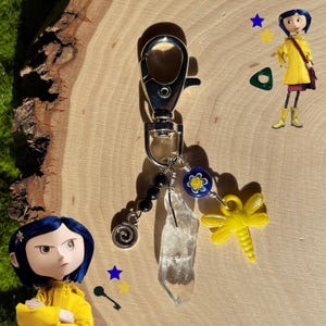 May include: A silver-toned keychain with a lobster clasp, adorned with black beads, a clear crystal, a yellow dragonfly charm, and a blue and yellow flower bead. The keychain is on a wooden surface.