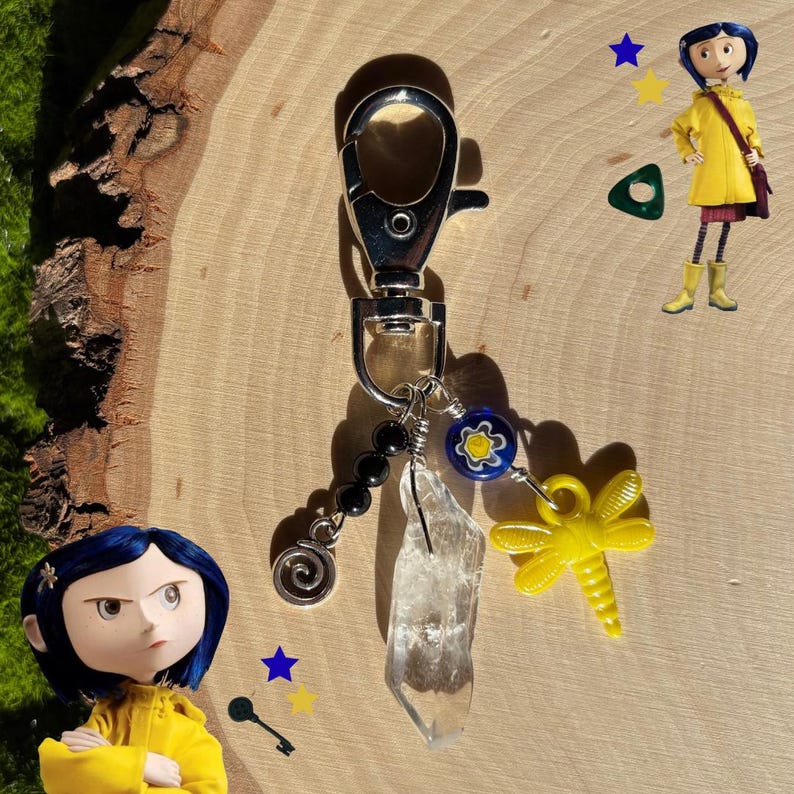 May include: A silver-toned keychain with a lobster clasp, adorned with black beads, a clear crystal, a yellow dragonfly charm, and a blue and yellow flower bead. The keychain is on a wooden surface.