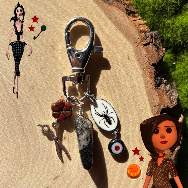 May include: Silver keychain with multiple charms, including a spider, a four-leaf clover, and a black and gray stone. The keychain also features small scissors and a red and black bead. The background is a wooden surface.