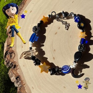 K&ouml;nnte beinhalten: Ein farbenfrohes Perlenarmband mit Stern-, Mond- und Wirbelanh&auml;ngern. Das Armband enth&auml;lt schwarze, blaue und gelbe Perlen sowie einen silberfarbenen Verschluss. Im Hintergrund befindet sich eine Figur eines Charakters in einem gelben Mantel.