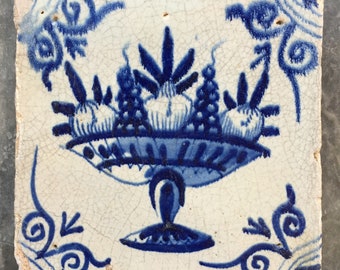 May include: A blue and white ceramic tile with a hand-painted design of a fruit bowl filled with grapes and other fruits. The bowl is on a pedestal and surrounded by decorative swirls.
