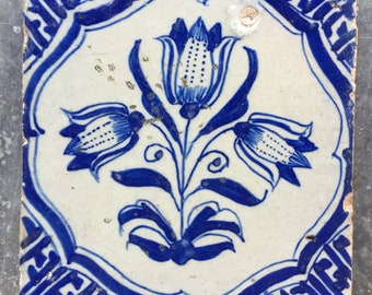 May include: A blue and white Delft tile with a floral design. The tile features three stylized flowers with blue and white petals and leaves. The tile is surrounded by a blue and white border.