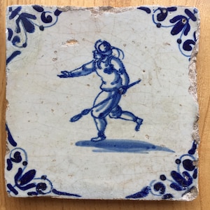 Könnte beinhalten: Eine blaue und weiße Delft-Kachel mit der Darstellung einer laufenden Figur mit einem Stock in der Hand. Die Kachel hat einen dekorativen Rand mit floralen Motiven.