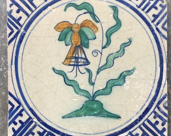May include: A vintage Dutch tile with a blue and white border and a central circle featuring a hand-painted floral design. The flower is yellow, orange, and green with a green stem and leaves.
