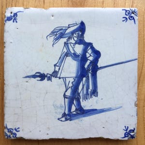 Può includere: Una piastrella di Delft blu e bianca che raffigura un soldato con un cappello piumato che tiene una lancia. La piastrella ha una glassa craquelé e un bordo decorativo.