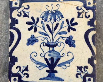 May include: A blue and white ceramic tile with a floral design. The tile features a vase with a floral arrangement, including grapes, and a blue and white border.