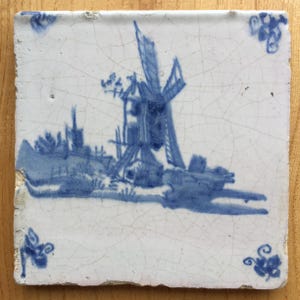 Può includere: Una piastrella di Delft blu e bianca che raffigura un mulino a vento in un paesaggio. La piastrella ha uno sfondo bianco con un mulino a vento blu, alberi e una recinzione. La piastrella è incrinata e ha un aspetto usurato.