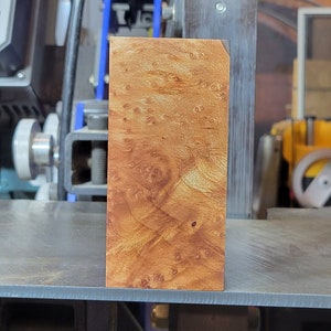 May include: A close-up of a block of wood with a unique, intricate grain pattern. The wood is a rich, warm brown color with streaks of yellow and orange. The block is sitting on a metal surface.