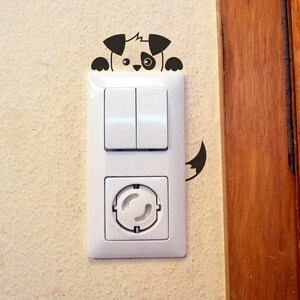 Könnte beinhalten: Eine weiße Steckdose und ein Lichtschalter mit einem schwarzen und weißen Hundesticker, der hinter dem Schalter hervorlugt.