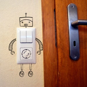 Könnte beinhalten: Ein weißer Lichtschalter und eine Steckdose mit einem schwarz-weißen Roboter, der an die Wand gezeichnet ist.