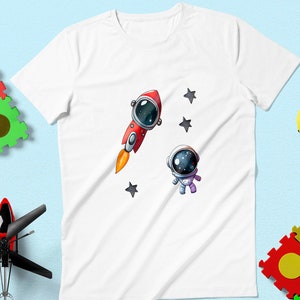 Könnte beinhalten: Weißes T-Shirt mit einer rot-weißen Rakete und einem blauen Astronauten mit schwarzem Helm und Sternen auf weißem Hintergrund.