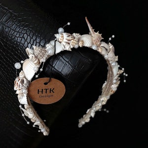 May include: A white headband decorated with seashells and pearls. The headband has a brown tag attached to it that says "HTK Design".