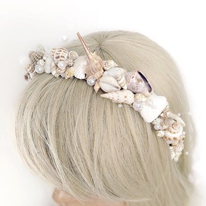 May include: A white seashell crown headband with pearl accents. The crown is made of various sizes and shapes of seashells.