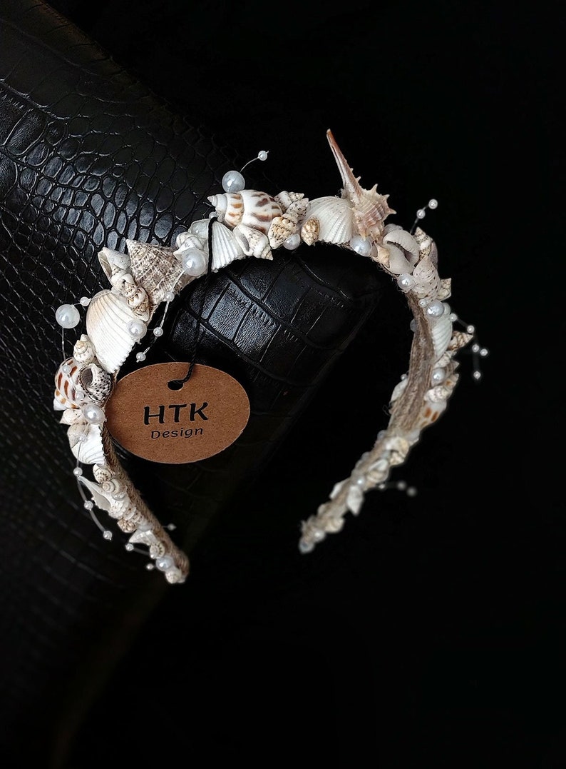 May include: A white headband decorated with seashells and pearls. The headband has a brown tag attached to it that says "HTK Design".