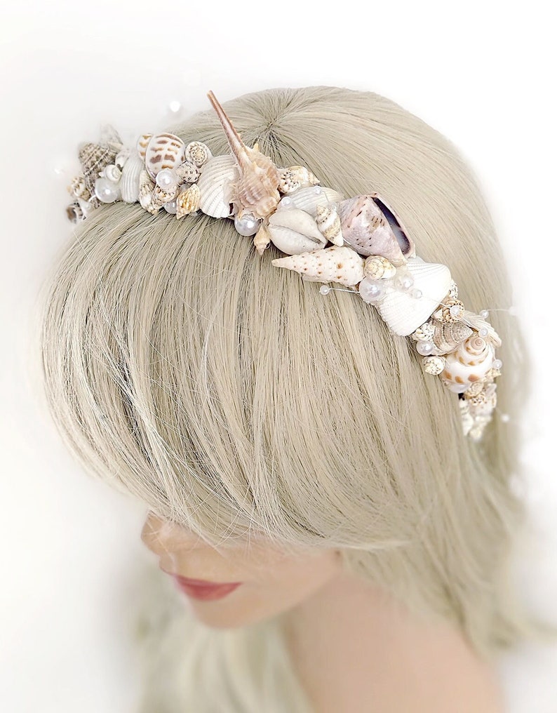 May include: A white seashell crown headband with pearl accents. The crown is made of various sizes and shapes of seashells.