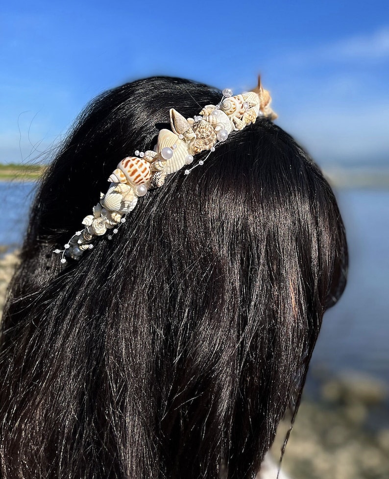May include: A white seashell headband with pearls and small seashells. The headband is worn on a person with long black hair.