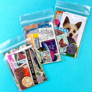 May include: Three clear plastic bags filled with assorted vintage ephemera, including stamps, maps, and decorative paper. One bag features a dog image. The bags are arranged on a bright blue surface, creating a colorful and organized display.