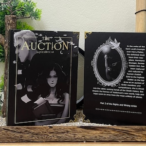 Könnte beinhalten: Zwei schwarze Bücher mit dem Titel The Auction von LovesBitca8. Das Cover zeigt eine dunkle Illustration von zwei Figuren. Die Rückseite zeigt einen weißen ovalen Rahmen mit einer Hand, die ein Licht hält, und Text zur Handlung.