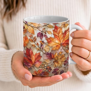 May include: A ceramic mug featuring a vibrant mosaic-style pattern of autumn leaves in shades of orange, red, yellow, and brown, interspersed with small clusters of berries against a light, textured background with a white interior and handle.