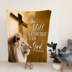 Puede incluir: Una manta de color marr&oacute;n y dorado con un le&oacute;n, una cruz y el texto "Be still & know that I am God Psalm 46:10".