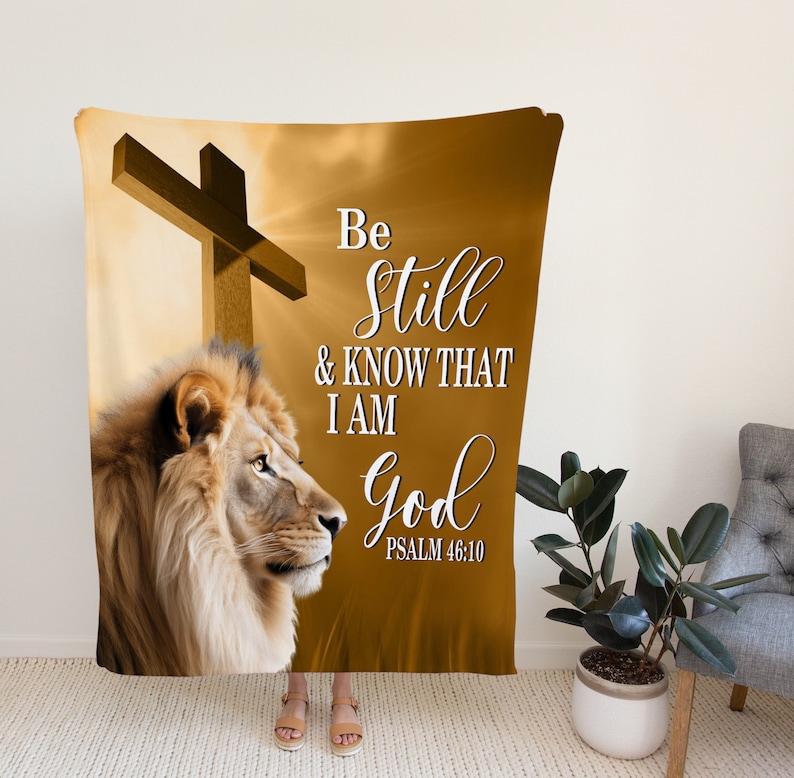 Puede incluir: Una manta de color marr&oacute;n y dorado con un le&oacute;n, una cruz y el texto "Be still & know that I am God Psalm 46:10".