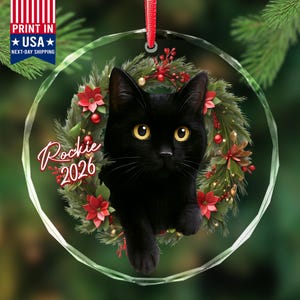 May include: A circular glass ornament featuring a black cat with yellow eyes centered within a festive wreath of green pine needles, red poinsettias, and small red berries. The text Rockie 2026 is printed in white script and block letters.