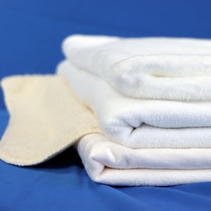 Puede incluir: Una pila de tres mantas de felpa blancas, una de color crema m&aacute;s claro, sobre un fondo azul.