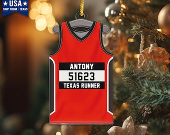 May include: Red and black running jersey ornament with the text "ANTONY 51623 TEXAS RUNNER." The ornament is hanging from a gold string. The background is a blurred Christmas tree with ornaments.