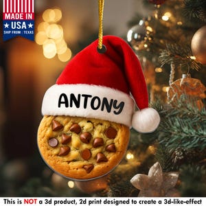 May include: A Christmas ornament shaped like a chocolate chip cookie, topped with a red Santa hat. The hat has the name "ANTONY" in black letters. The ornament is hanging from a gold string. The text "MADE IN USA" is visible.