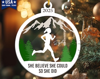 May include: A white running-themed Christmas ornament with a gold string. The ornament features a runner silhouette, mountains, and evergreen trees. Text includes "2025", "Sophia", and "SHE BELIEVE SHE COULD SO SHE DID".