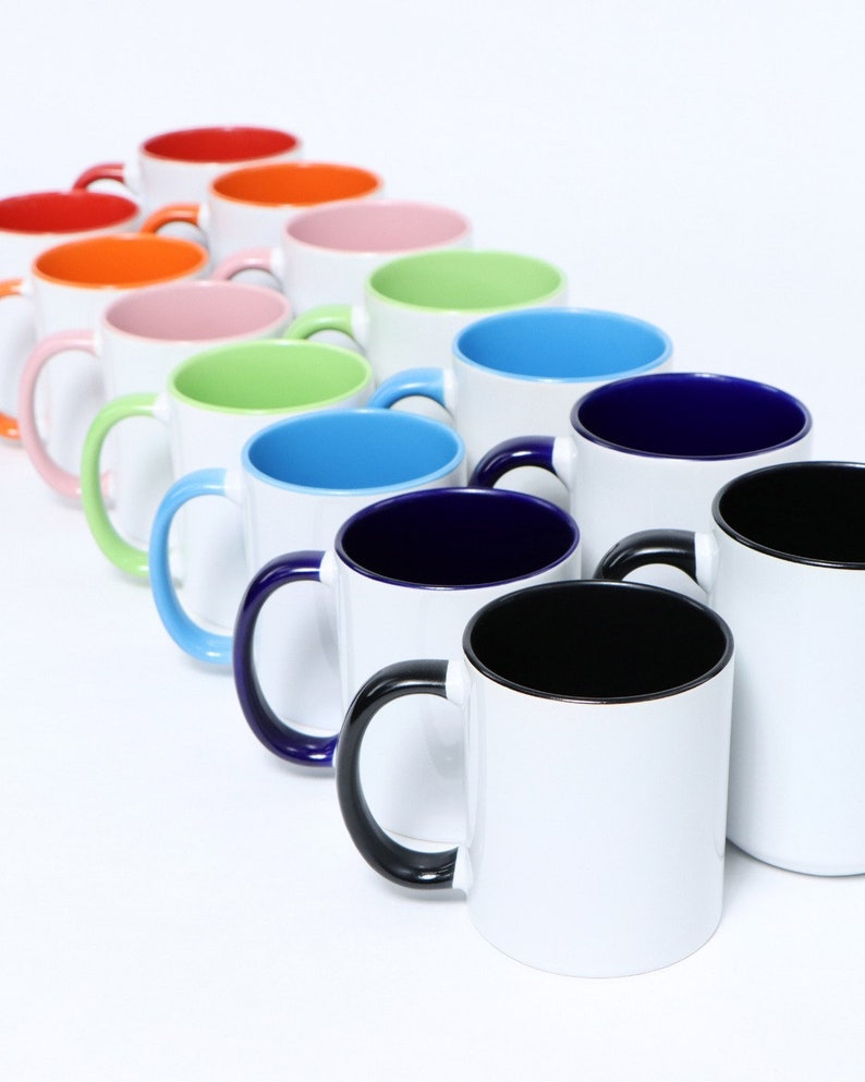 K&ouml;nnte beinhalten: Eine Reihe von 12 wei&szlig;en Keramikbechern mit farbigen Innenr&auml;umen. Die Becher haben schwarze Henkel und sind in einer Regenbogenfarbenfolge von Rot bis Schwarz angeordnet.