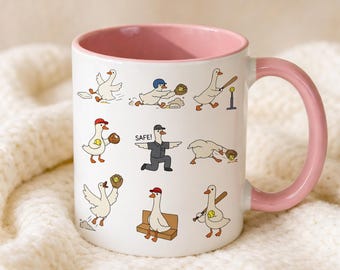 May include: White ceramic mug with a pink interior and handle. The mug features cartoon geese playing baseball. The geese are depicted in various baseball-related poses, with some wearing baseball caps. The word "SAFE!" is printed on the mug.