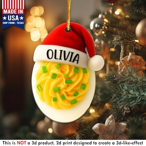 May include: A deviled egg Christmas ornament with a Santa hat. The ornament is white, yellow, red, and green. The name "OLIVIA" is printed on the hat. The ornament is made in the USA and ships from Texas.