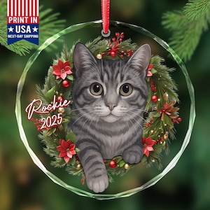 May include: A clear, round Christmas ornament featuring a gray tabby cat within a wreath of greenery, red berries, and poinsettias. The ornament has the name "Rockie" and the year "2025" in red script. A red ribbon is attached for hanging.