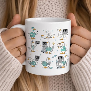 May include: White ceramic mug featuring cartoon illustrations of geese in medical attire. The geese are depicted as doctors and nurses, using medical equipment. The mug has a handle and is held by a person.