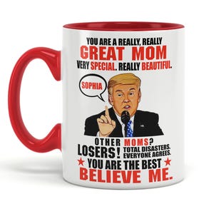 May include: White ceramic coffee mug with a red rim. The mug features a cartoon image of a man with a red tie and a white shirt, speaking into a microphone. The text on the mug reads "You are a really, really Great Mom Very Special. Really Beautiful. Sophia Other Moms? Losers! Total Disasters. Everyone agrees. You are the best Believe Me."