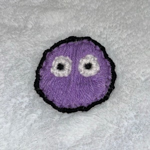 May include: A handmade, embroidered purple blob with white and black eyes. The blob is outlined in black thread, giving it a defined shape. The craft item is made of yarn and is likely a patch or applique.