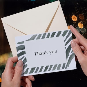 Puede incluir: Una tarjeta blanca con las palabras "Thank you" en una fuente sencilla, enmarcada por un diseño de rayas de cebra estilo acuarela en tonos grises y verdes. La tarjeta se sostiene frente a un sobre color crema.