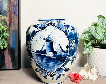 May include: A white ceramic vase with a blue and white Delft design featuring a windmill and a canal scene. The vase has a round body and a wide mouth.