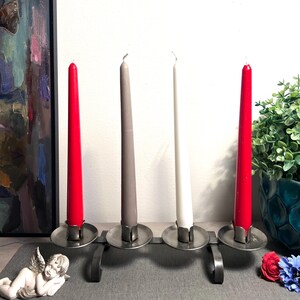 Könnte beinhalten: Ein Metallkerzenhalter mit vier Kerzen. Zwei rote, eine graue und eine weiße Kerze befinden sich in der Halterung. Der Halter steht auf einer grauen Oberfläche mit einer kleinen Engelsstatue und einem Gemälde im Hintergrund.