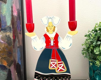 May include: A wooden candle holder in the shape of a woman in traditional attire, holding two red candles. The figure is painted with a red, white, blue, and yellow color scheme, with floral and geometric details.