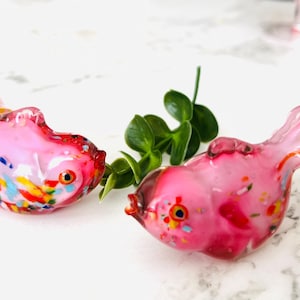 May include: Two pink glass goldfish figurines with colorful flecks. The goldfish are facing each other and have a shiny, iridescent finish.