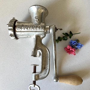 May include: A vintage silver-colored metal hand-crank meat grinder with a wooden handle. The grinder has a clamp to attach it to a surface and a crank to turn the grinder. The grinder has the word "HUSQVARNA" and the number "5" on it.