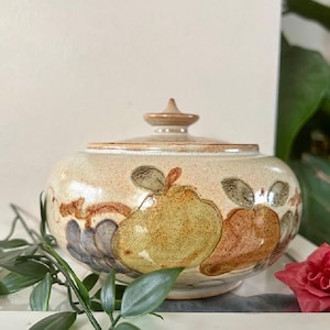 May include: A ceramic lidded jar with a cream-coloured base and a brown, yellow, and green floral design. The jar is decorated with a stylised floral pattern featuring pears and grapes.