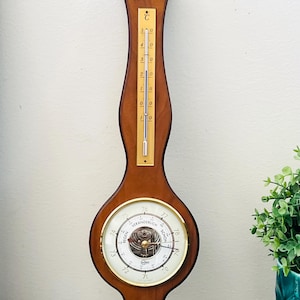 May include: A wall-mounted weather station featuring a wooden frame with a vertical thermometer, a central barometer with a white dial and gold-tone bezel, and a smaller hygrometer at the bottom. The barometer dial includes the text REGEN, VERANDERLICH, and SCHON.